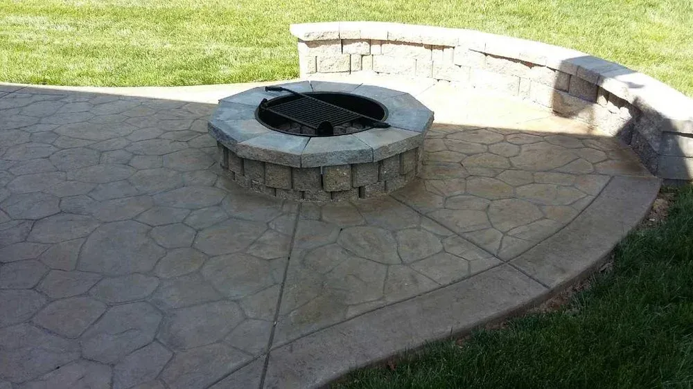 Round stone fire pit on a paved patio beside a curved stone bench and grass lawn.