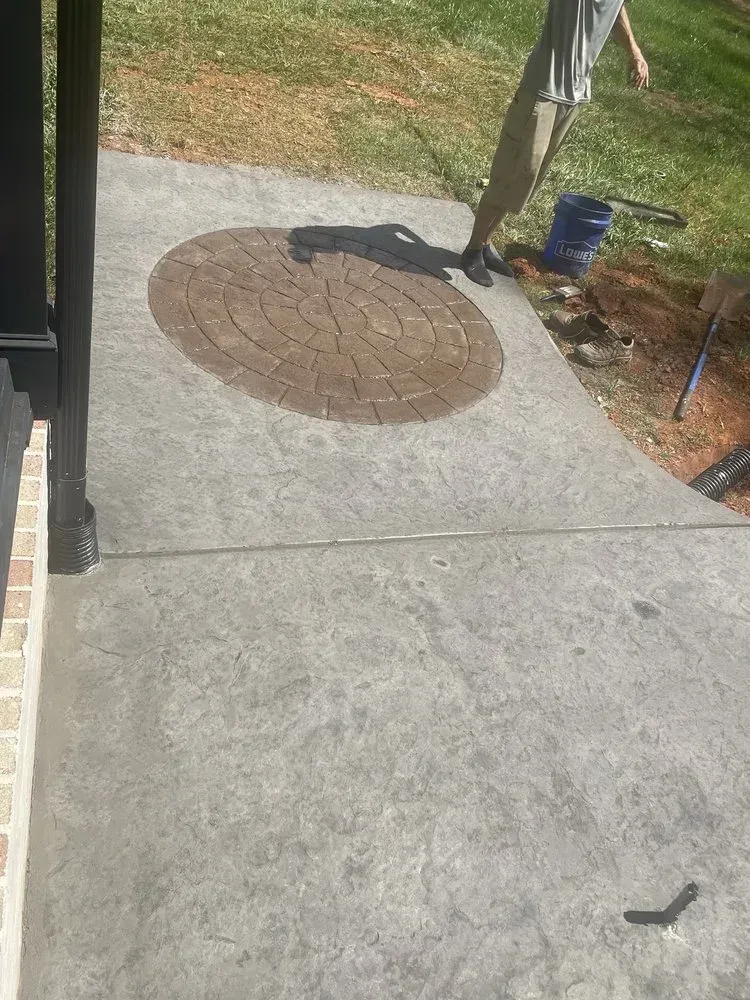Concrete porch with a round brown mat, doorway on left, and yard tools in the background.