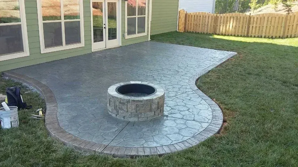 Backyard patio with curved stone fire pit and stamped concrete beside a lawn and house.