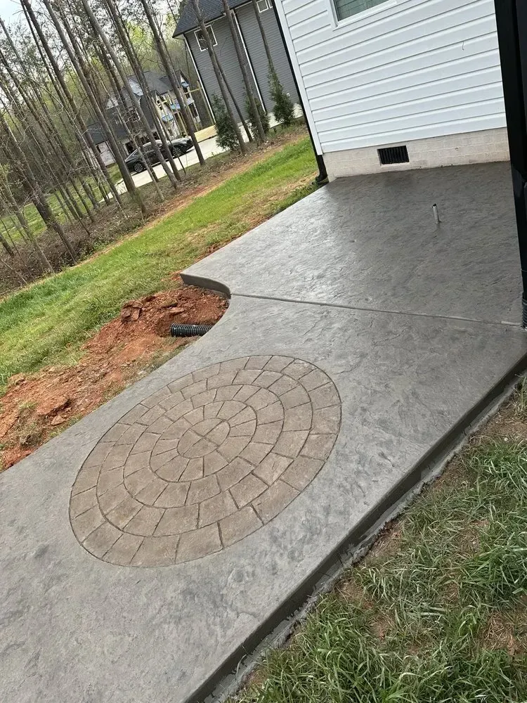 Concrete patio beside white house with circular brick inlay and unfinished edge near grass and trees