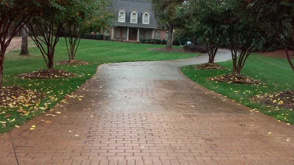 Brick driveway leading to a house through a green, tree-lined yard.