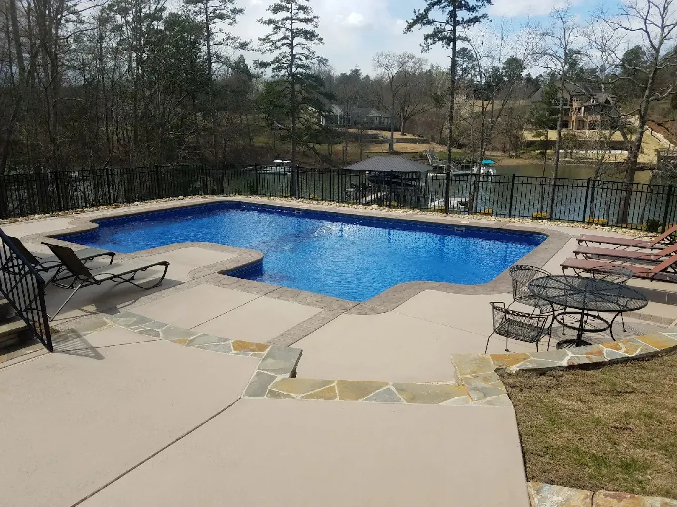 Backyard pool with blue water, stone patio, lounge chairs, and trees in the background
