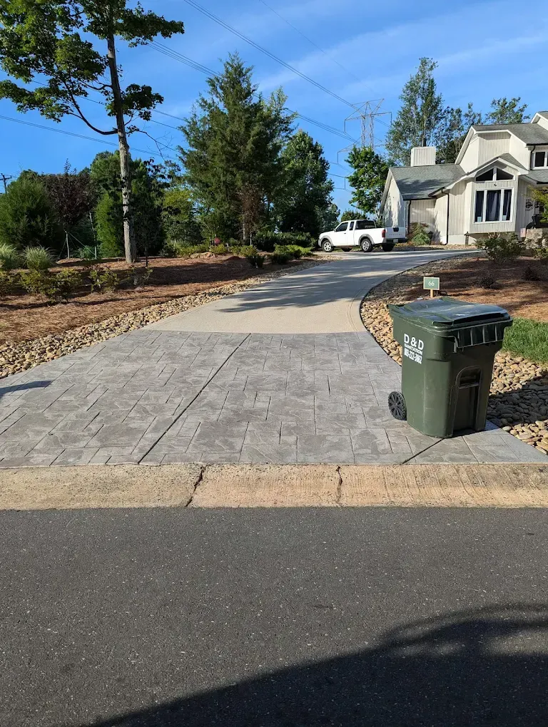 Residential driveway with a green trash bin, sidewalk, trees, and houses on a sunny day