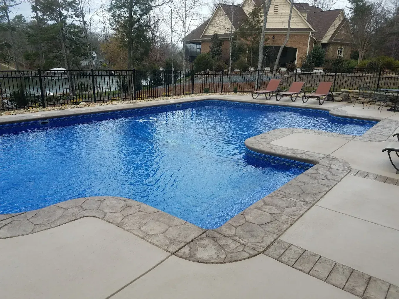 Blue swimming pool with stone deck, lounge chairs, and a small raised spa in a fenced backyard