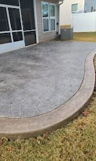 Stamped concrete patio beside a house, bordered by a curved concrete edge and small grass strip.