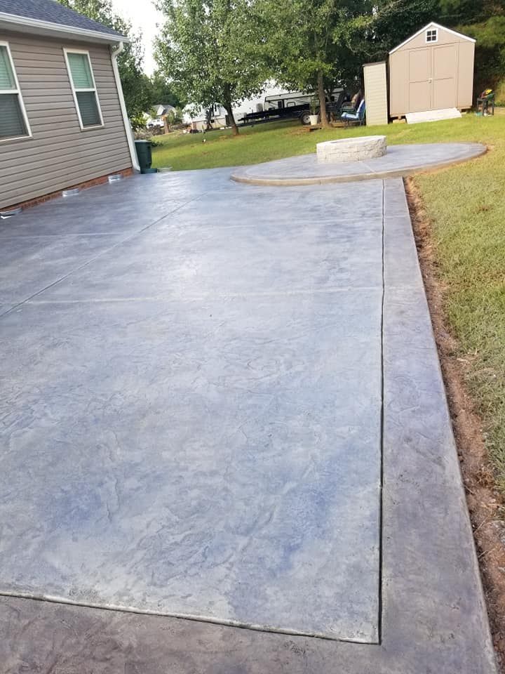 Freshly poured gray concrete patio beside a house, with a driveway and shed in the yard.
