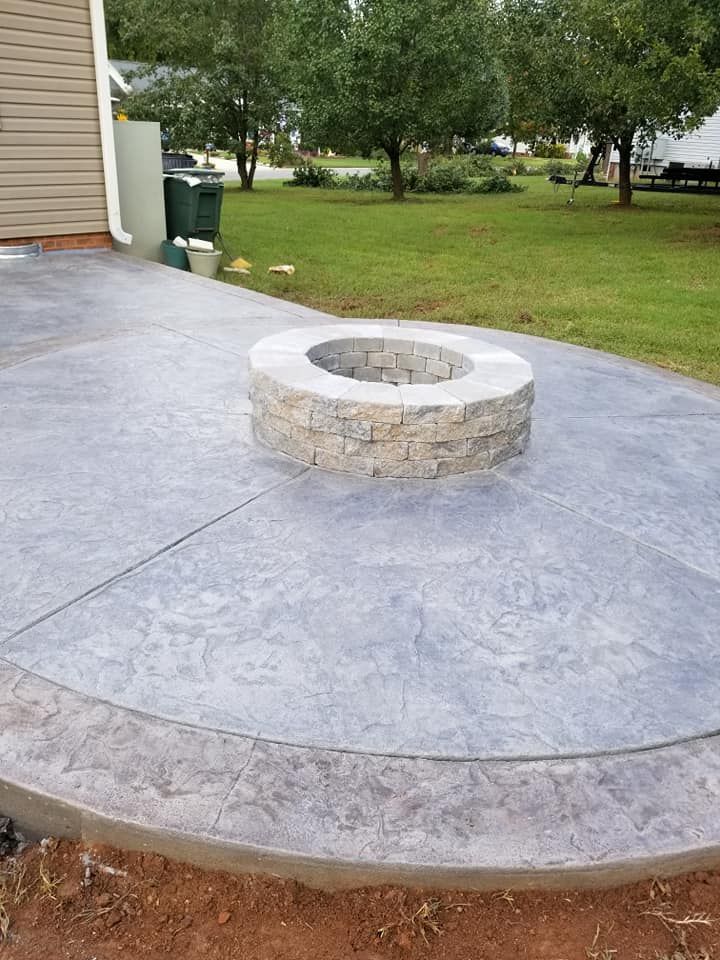 Stone fire pit on a circular concrete patio beside a house and grassy yard.