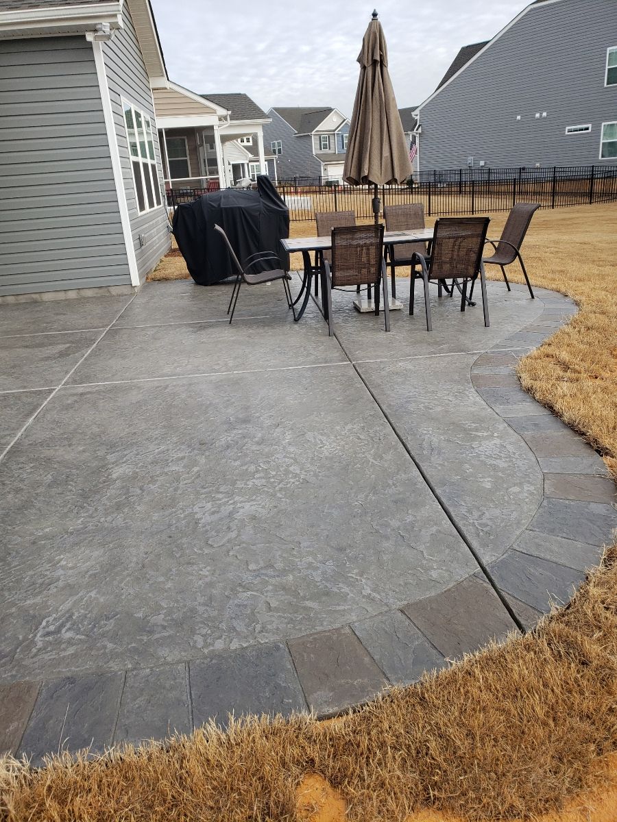 Backyard patio with curved stone edge, outdoor dining table, chairs, umbrella, and grill beside the house.