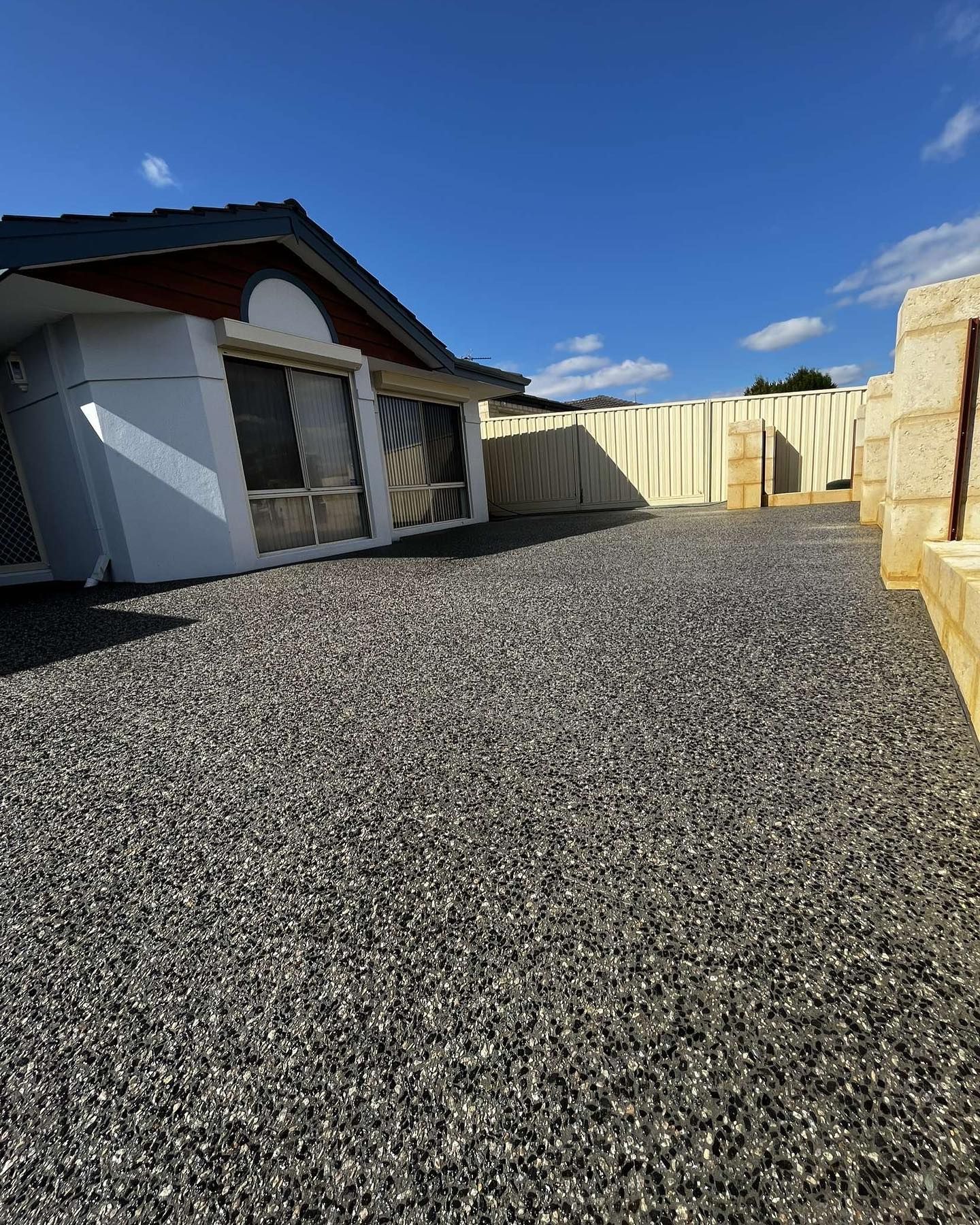 exposed aggregate driveway next to a house with a white exterior, blue sky overhead.