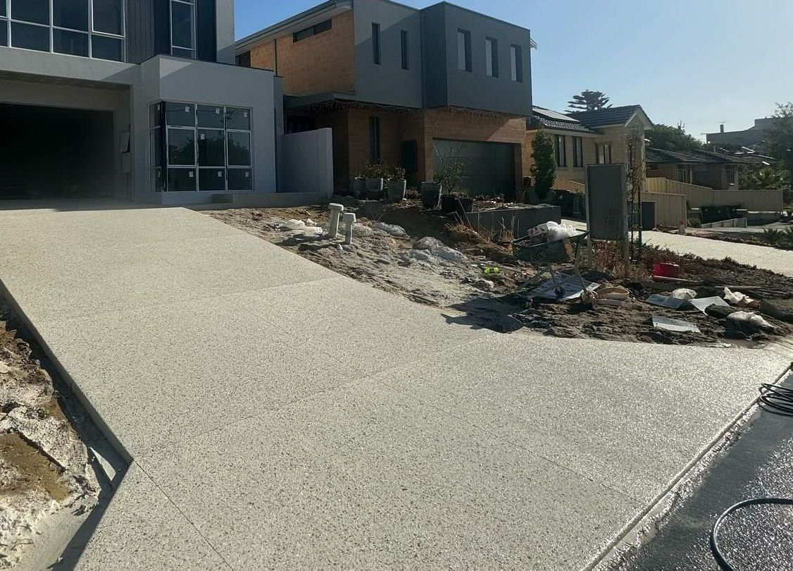 Light-colored concrete driveway leading to a modern house under construction, with exposed dirt and construction debris nearby.