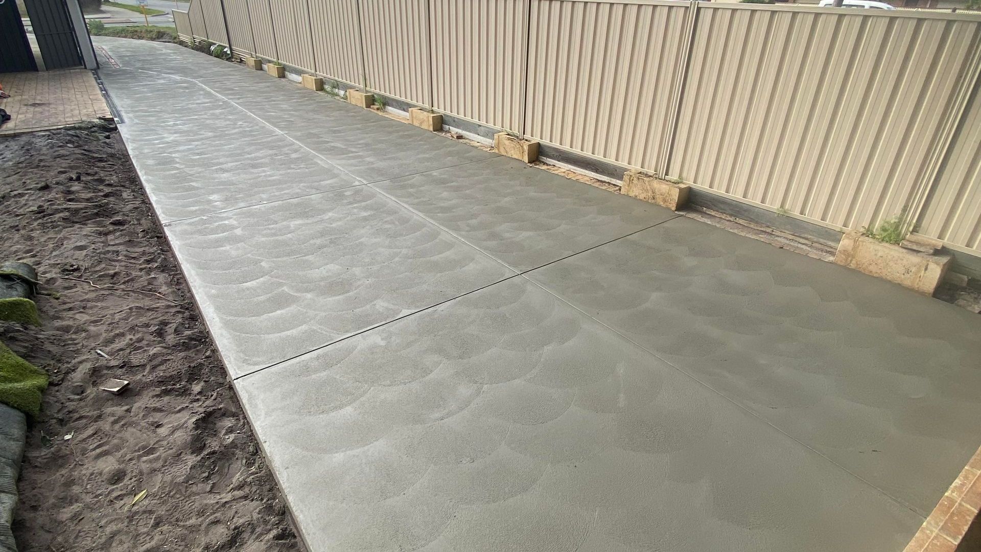 Freshly poured standard concrete driveway next to a colour bond fence and dirt.