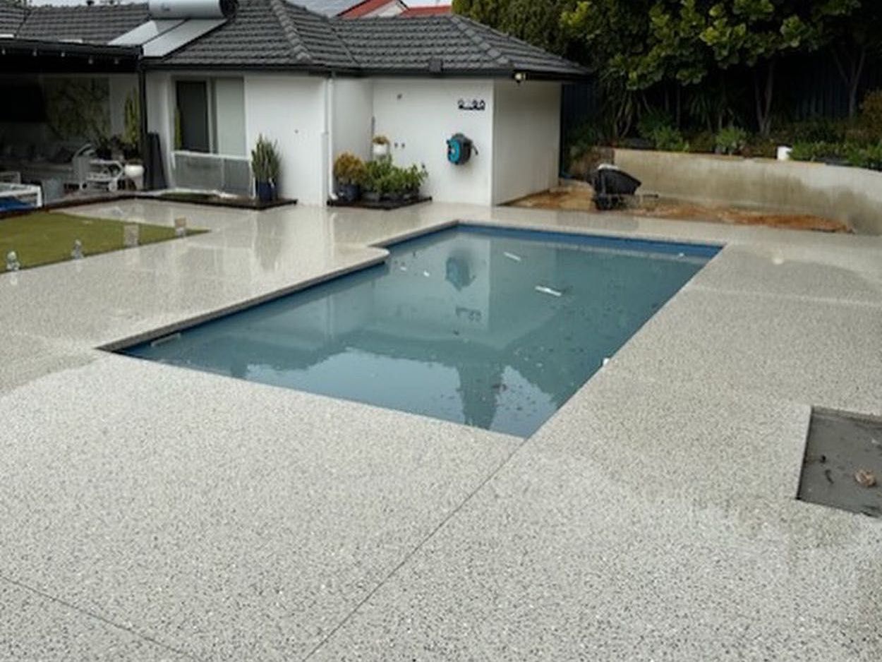 A rectangular swimming pool surrounded by a light-colored, speckled concrete pool surround in a residential backyard.