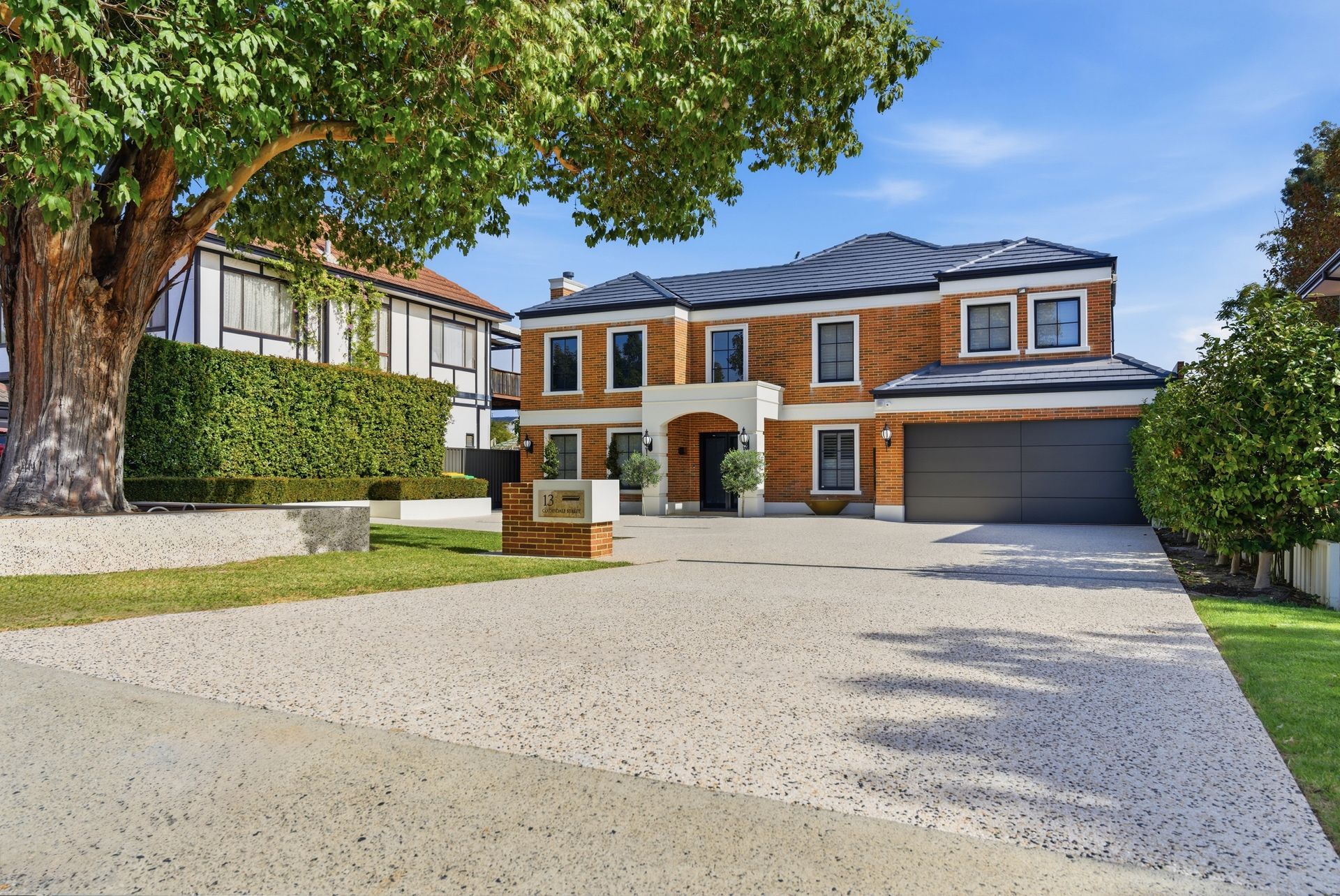 Large double garage exposed aggregate driveway to large two storey home with green gardens and hedges