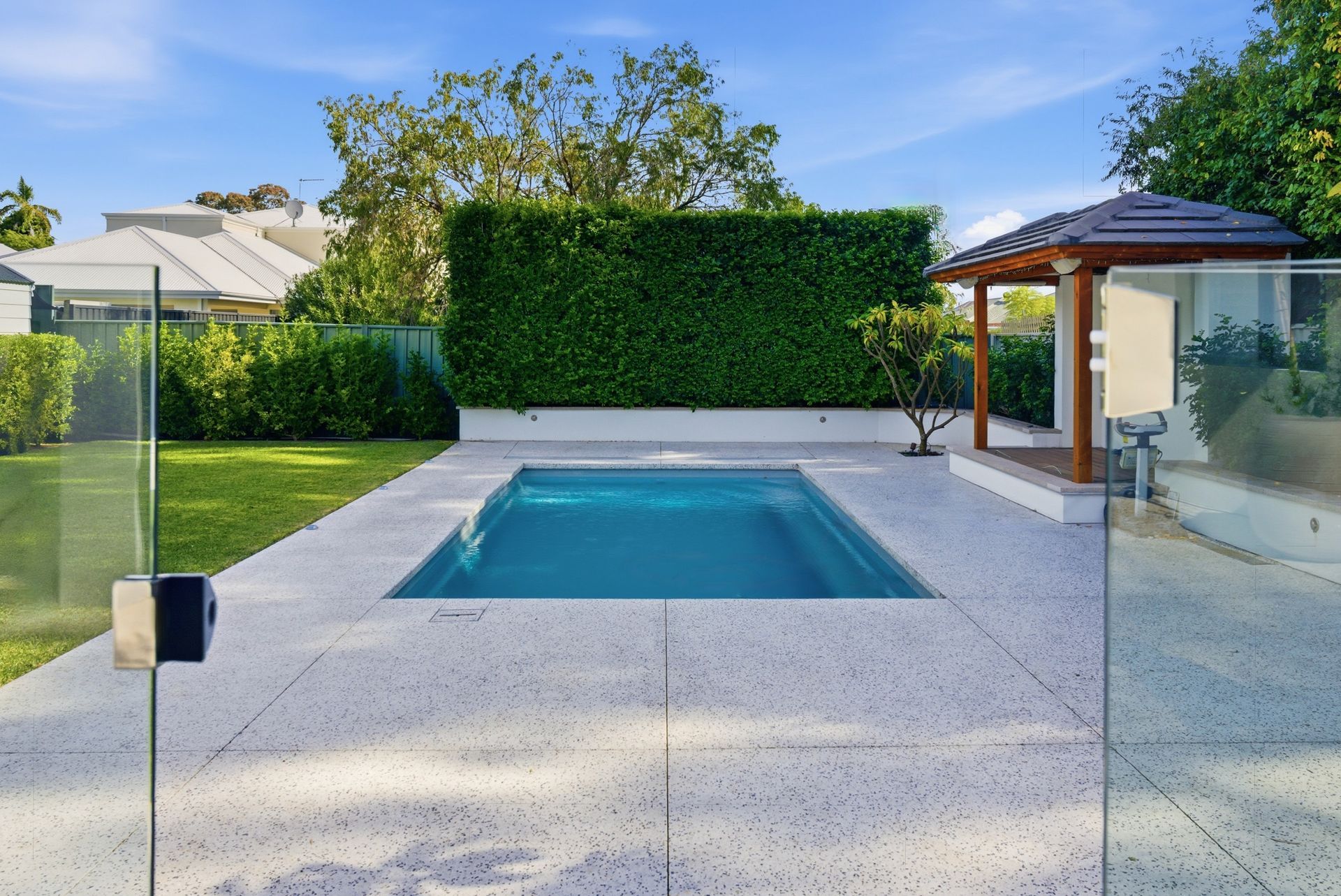 Modern backyard with a small rectangular pool, glass fence, and manicured hedges under a clear sky with a new honed concrete pool surrounds