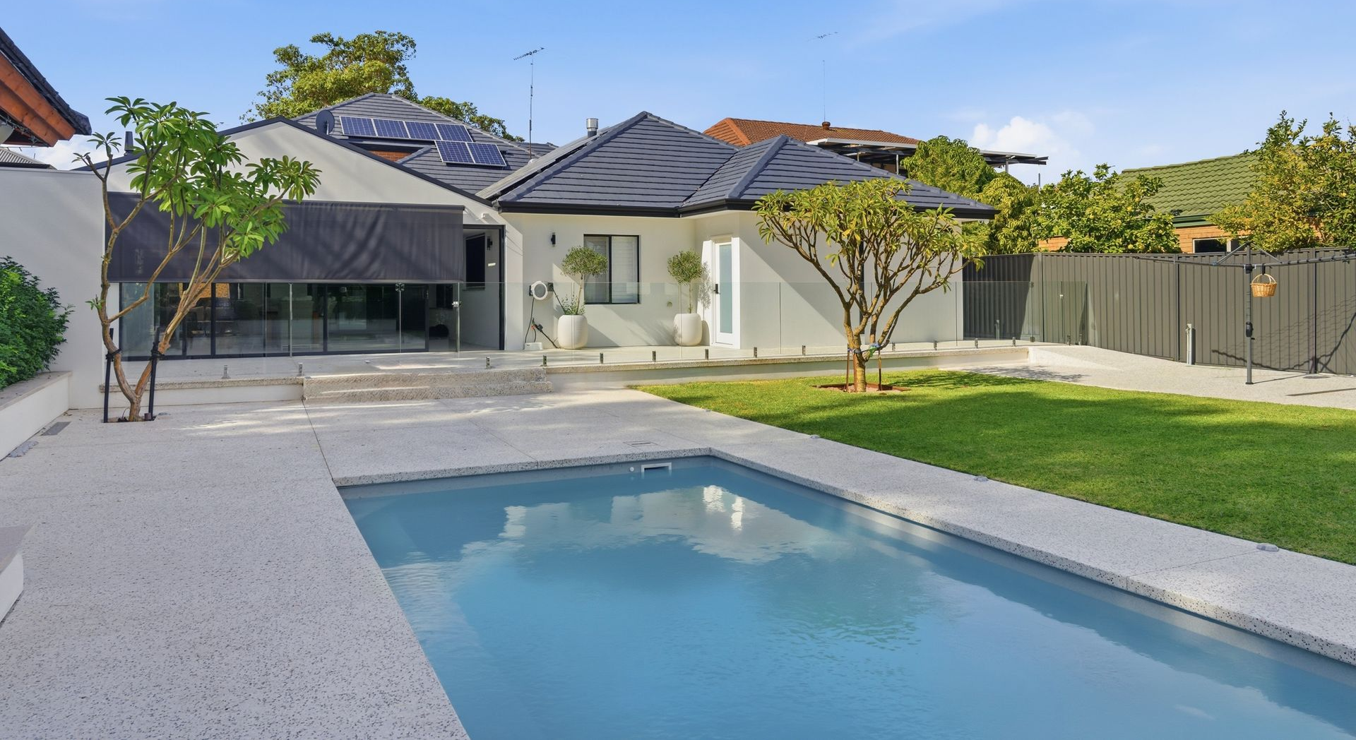 Backyard with a rectangular swimming pool surrounded by exposed aggregate concrete, Lush green lawn,