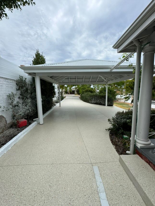 Driveway under a white carport with concrete path and driveway, vegetation on sides, and overcast sky.