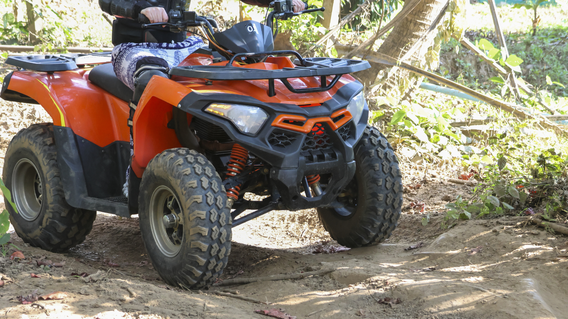 A bright orange ATV drives across a dirt path in a wooded area.