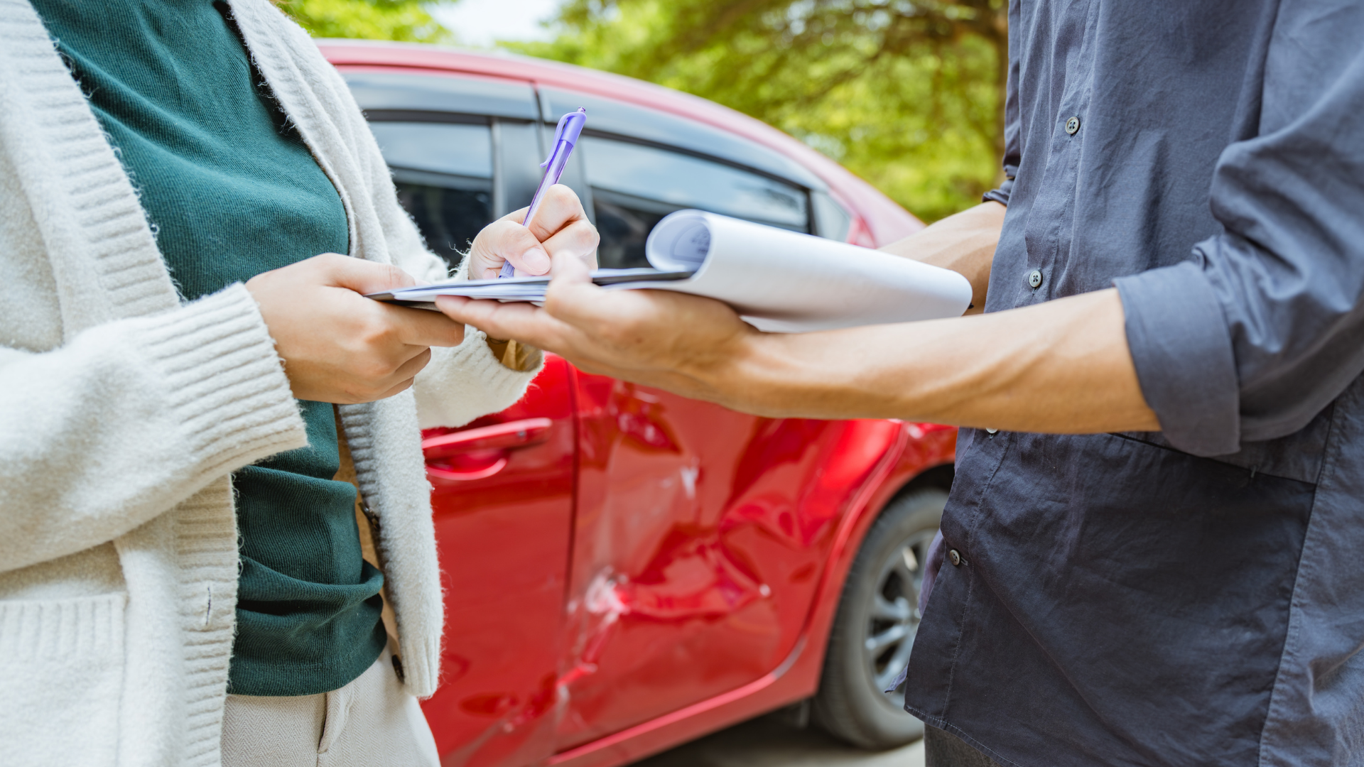 A person in a white cardigan signs a document held by another person in front of a red car with visible body damage.