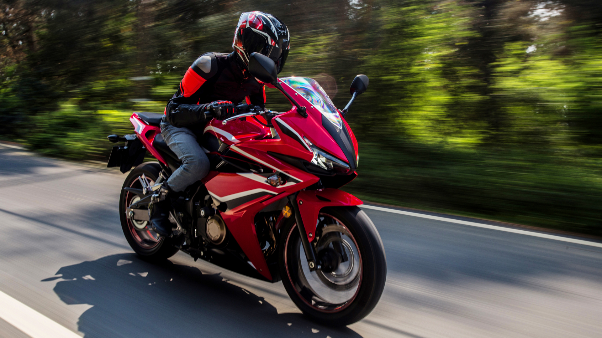 A motorcyclist in a black and red jacket rides a bright red sportbike along a tree-lined road with motion blur.