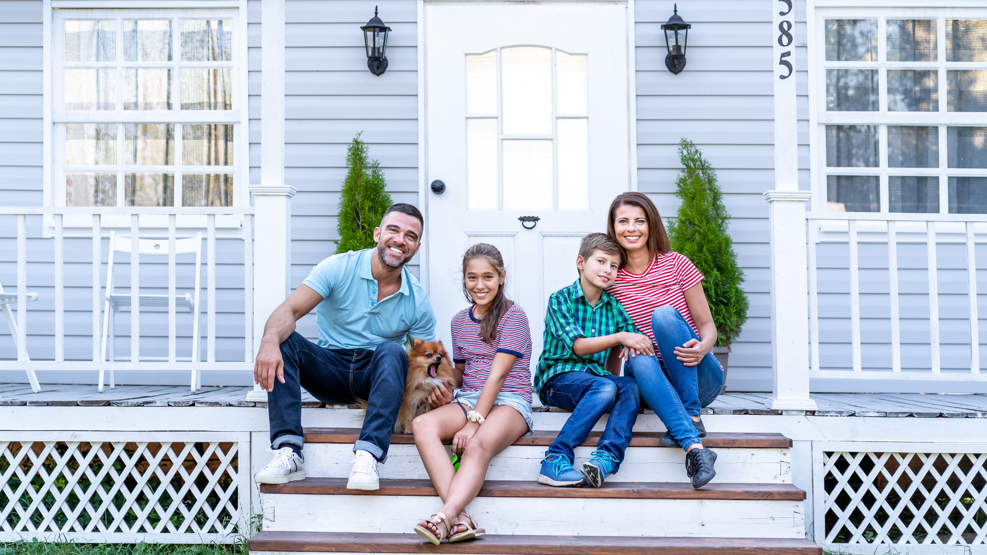 A family of four sits on the front porch steps of a light blue house, smiling together with a small dog.