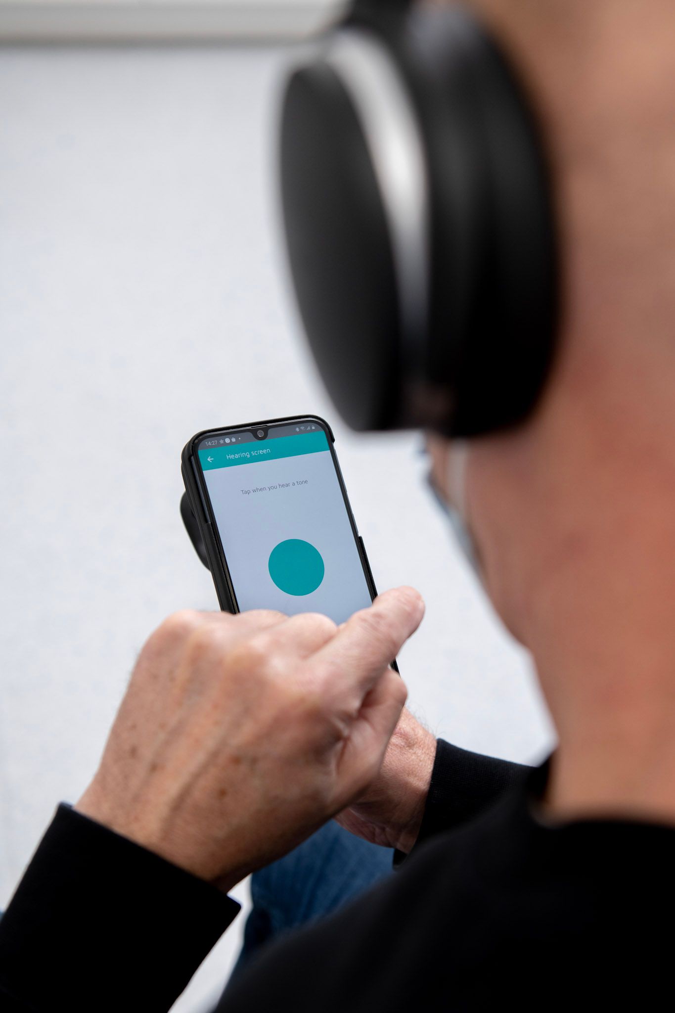 A person wearing large headphones interacts with a smartphone app displaying a teal button on a plain white background.