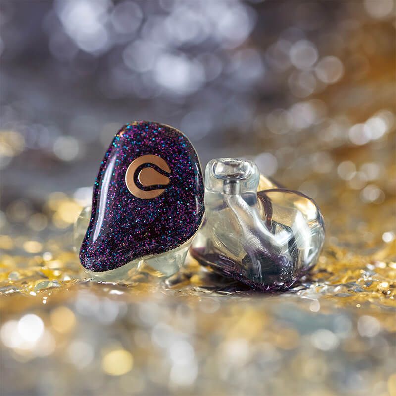 Two Cosmic Ears custom in-ear monitors with glittery purple faceplates and clear shells sitting on a glittering surface.