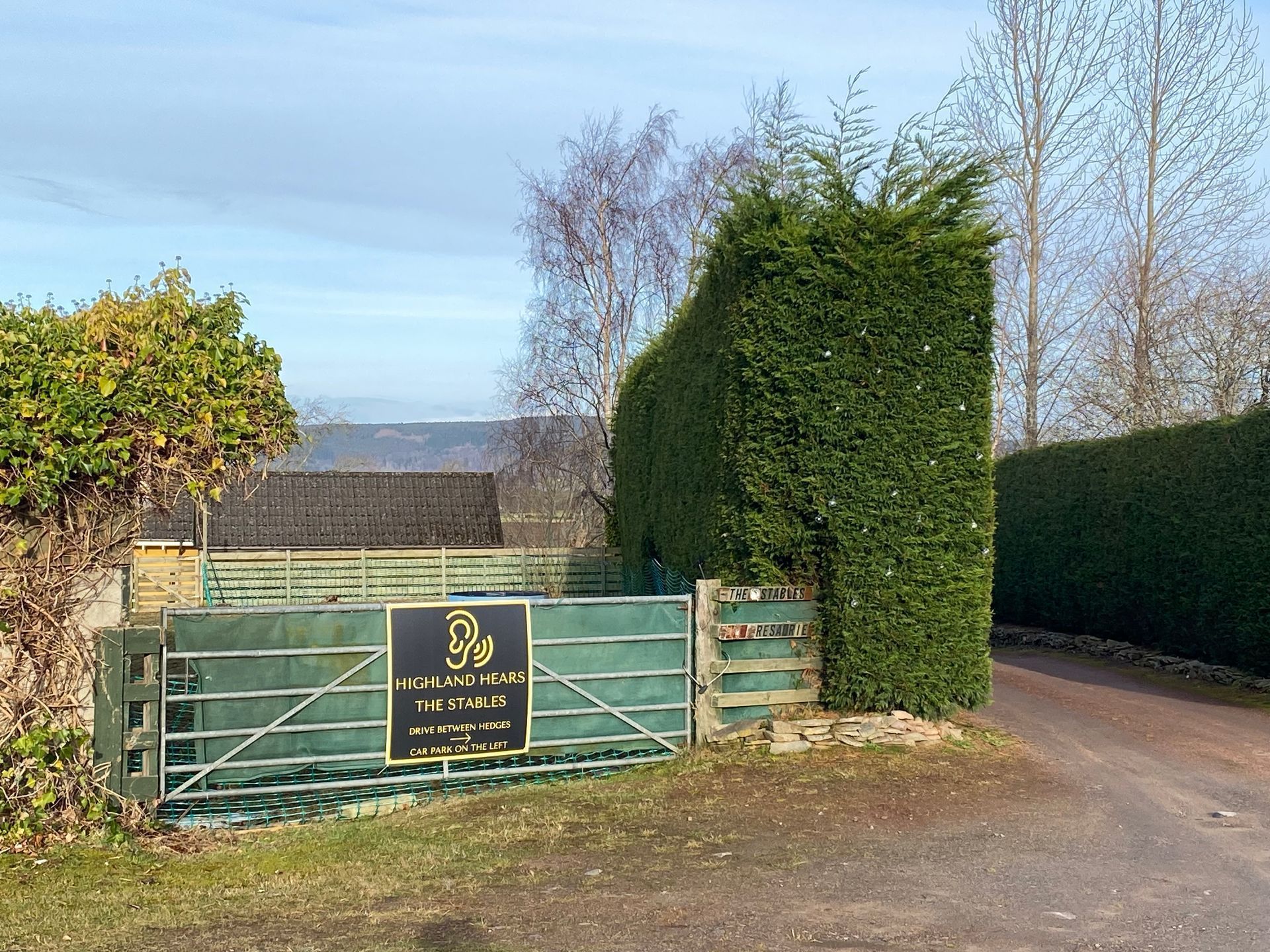 A green gated entrance to a property with a tall hedge and a black sign with gold lettering reading