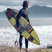 Surfer holding a colorful surfboard on a windy beach with waves and distant cliffs in the background
