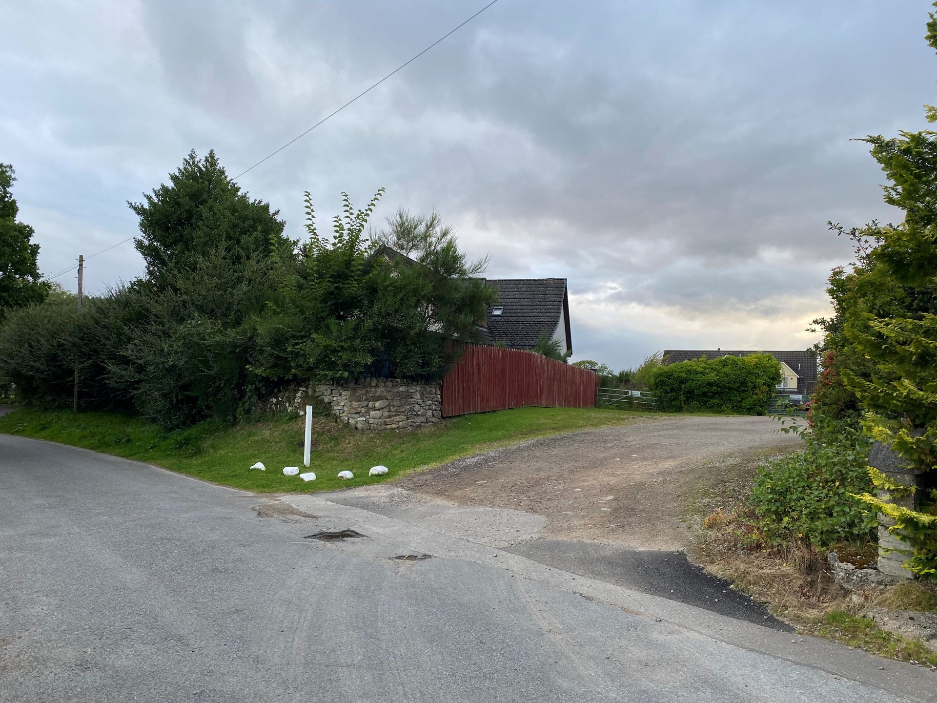 A gravel road turns right towards a house behind a stone wall and red wooden fence under a cloudy sky.