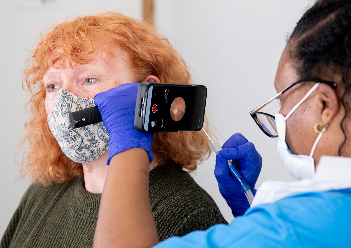 A clinician wearing blue gloves uses a digital otoscope attached to a smartphone to examine a patient's ear.