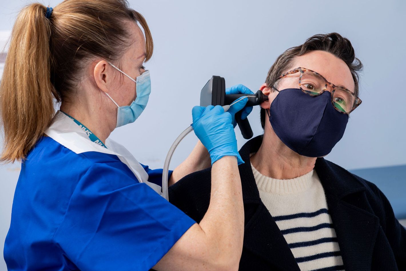 A healthcare professional in blue scrubs and a face mask examines a patient's ear with an otoscope in a clinical setting.