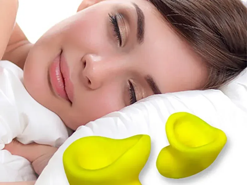 A person sleeping peacefully next to two small, yellow, cup-shaped silicone earplugs on a white pillow.