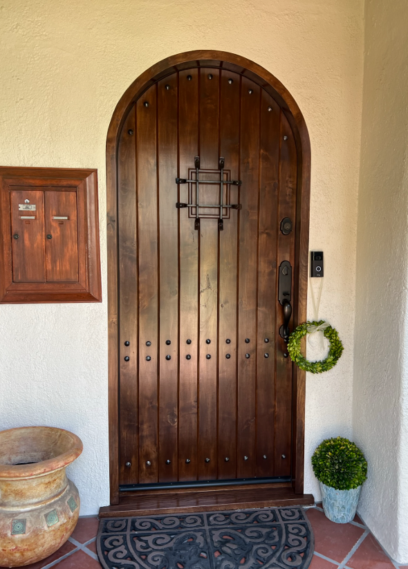 Wooden arched front door with wrought iron accents, mail slot, and greenery.