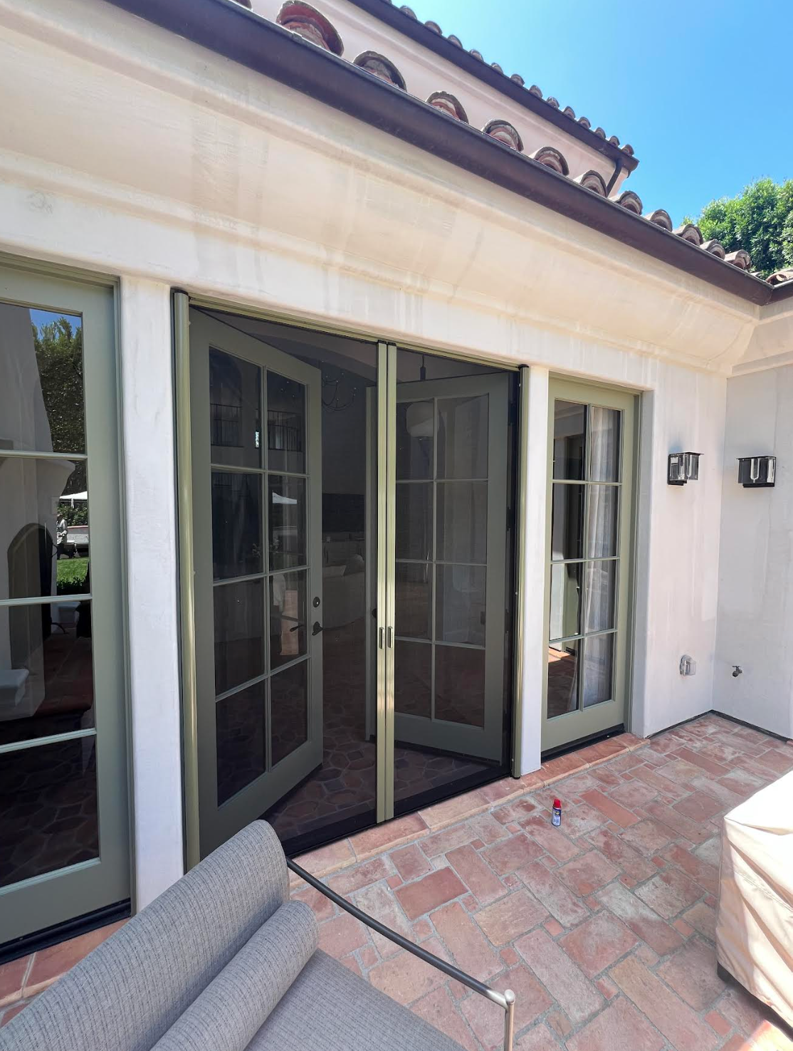 Outdoor patio with French doors, green frames, and screen doors. Brick patio, tan walls, blue sky.