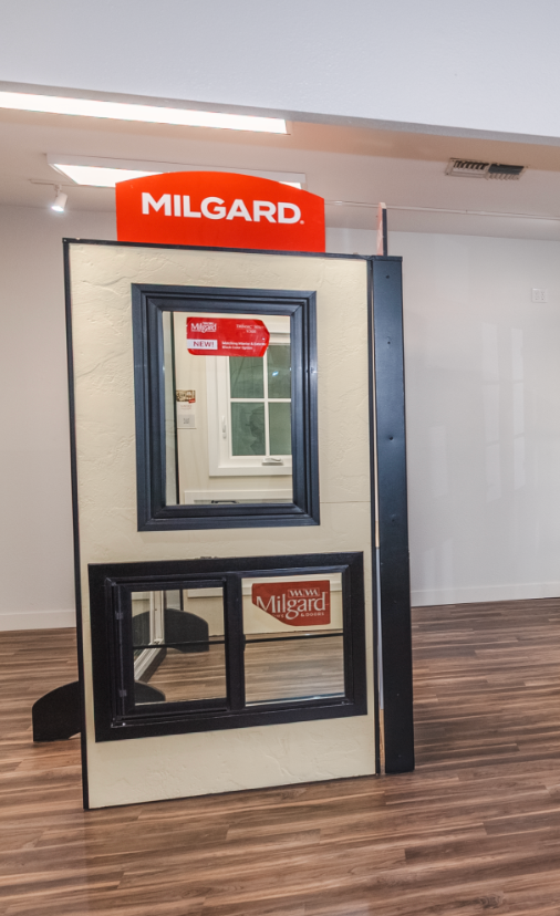 Display of Milgard windows, black frames, tan wall, wood flooring, orange sign.