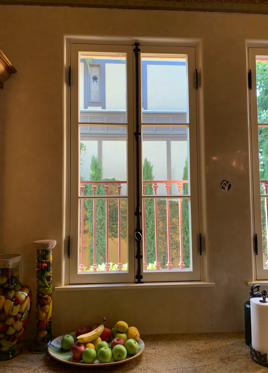 Window overlooking a courtyard with green trees, a red railing, and a plate of fruit on a counter.