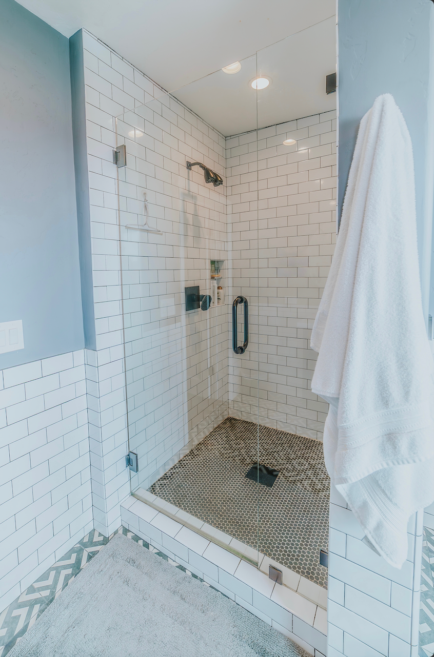 Shower with white brick tiles, glass door, and pebble floor. A white towel hangs on the wall.