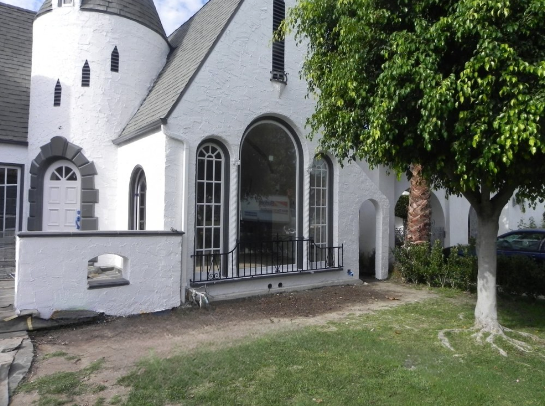 White church with arched windows and a gated entrance beside a large tree and grassy lawn