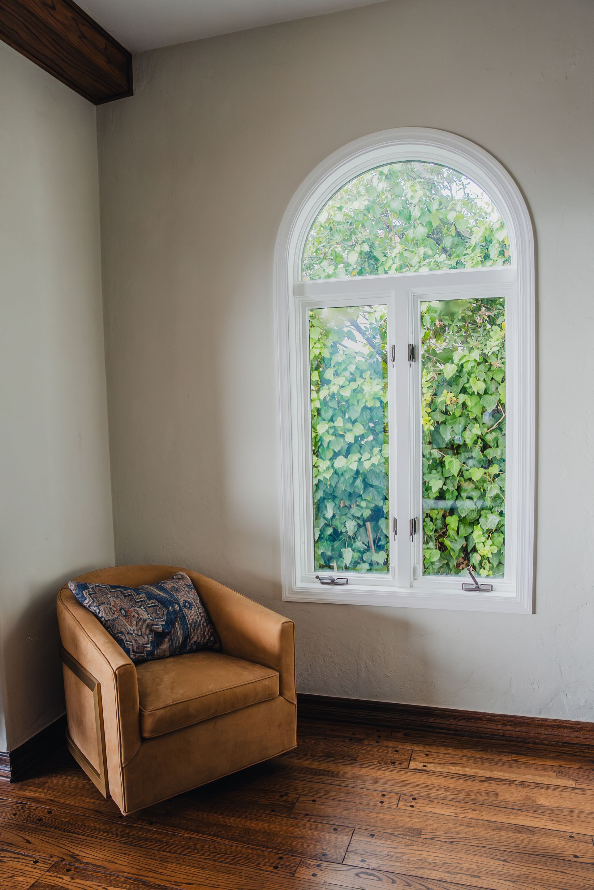 A cozy armchair with a patterned pillow sits by a tall arched window overlooking greenery in a corner of a room.