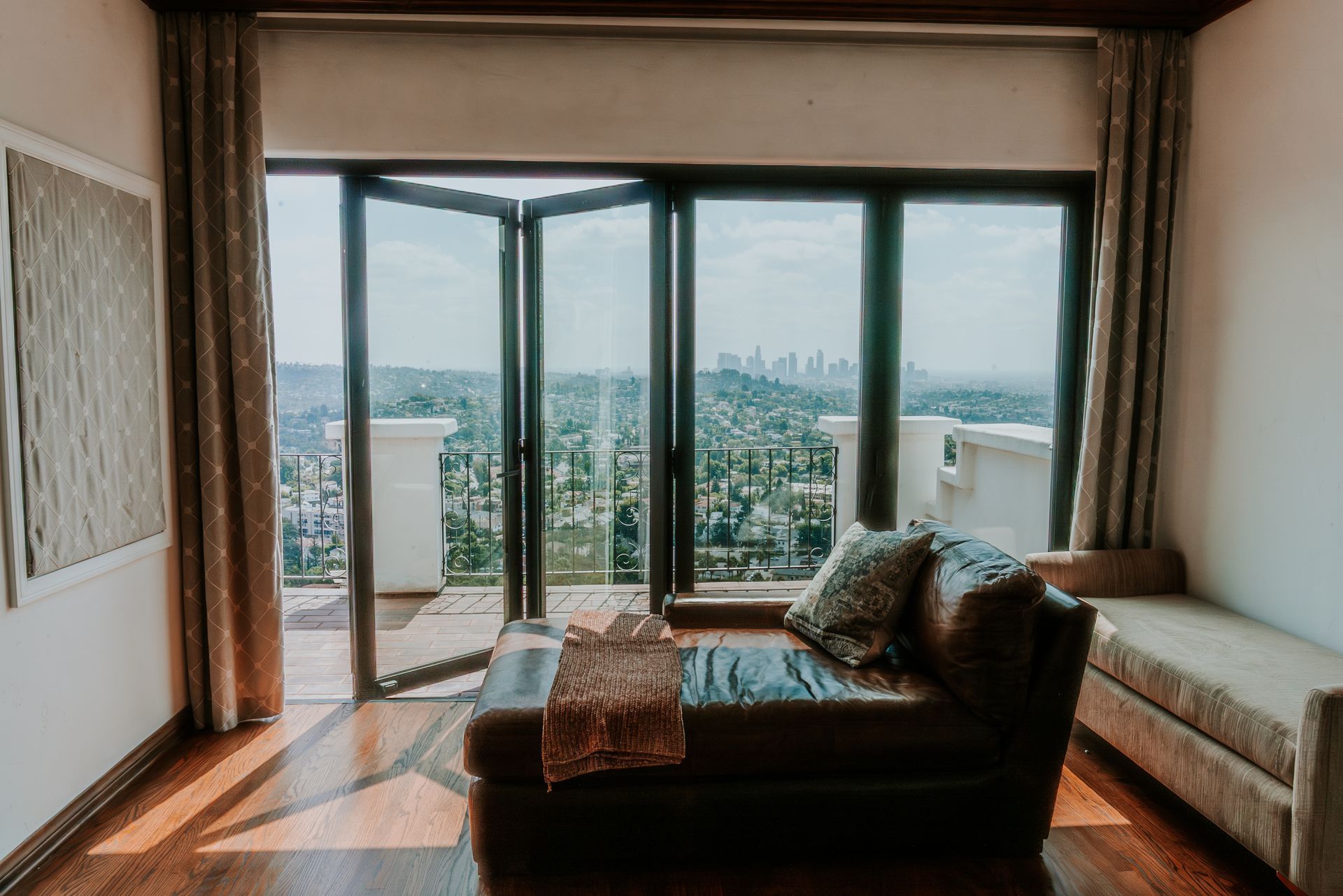 A brown leather chaise lounge sits before a large open glass door with a scenic city view.