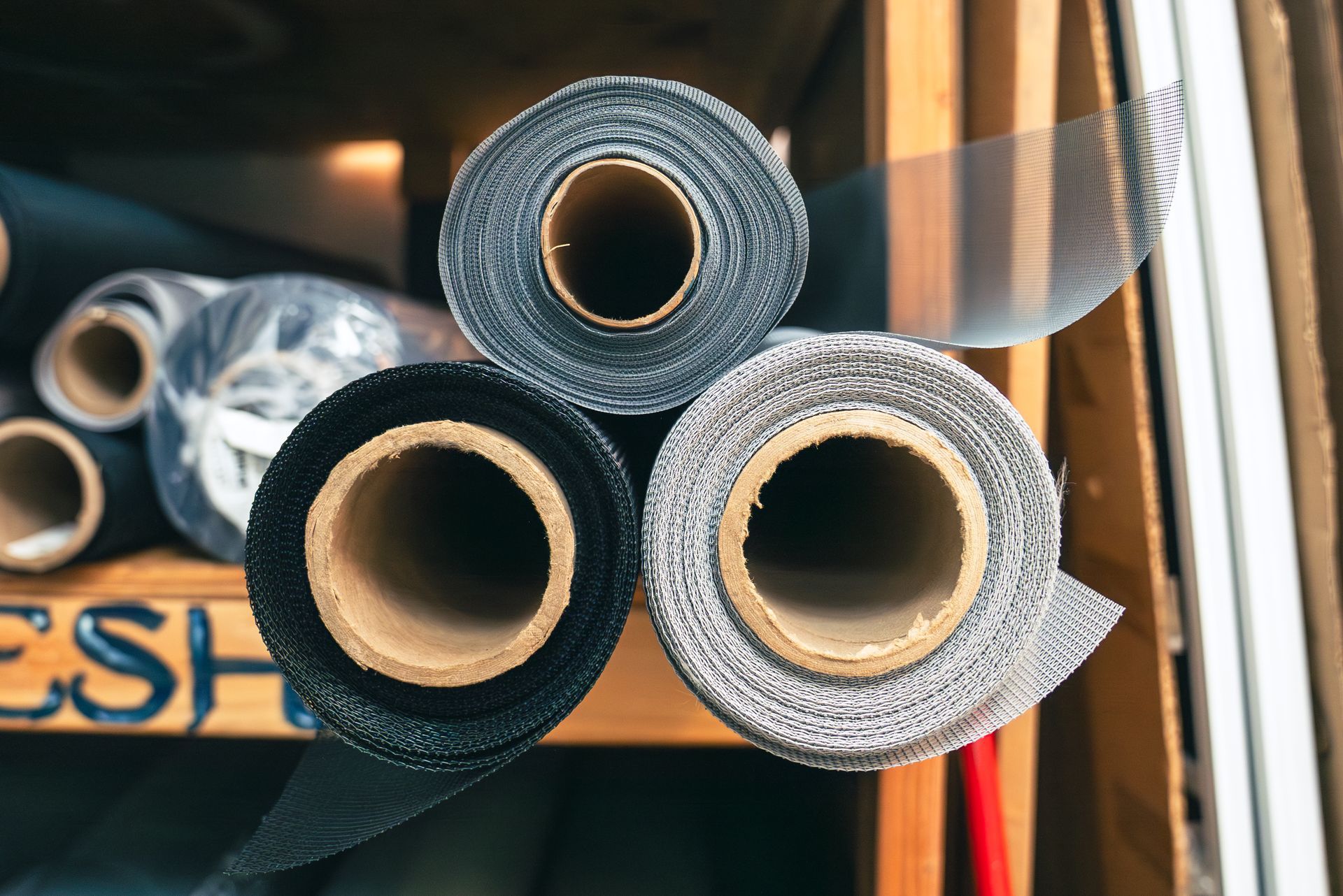 Three rolls of screen mesh, black, gray, and silver, stacked on a wooden shelf in a store.