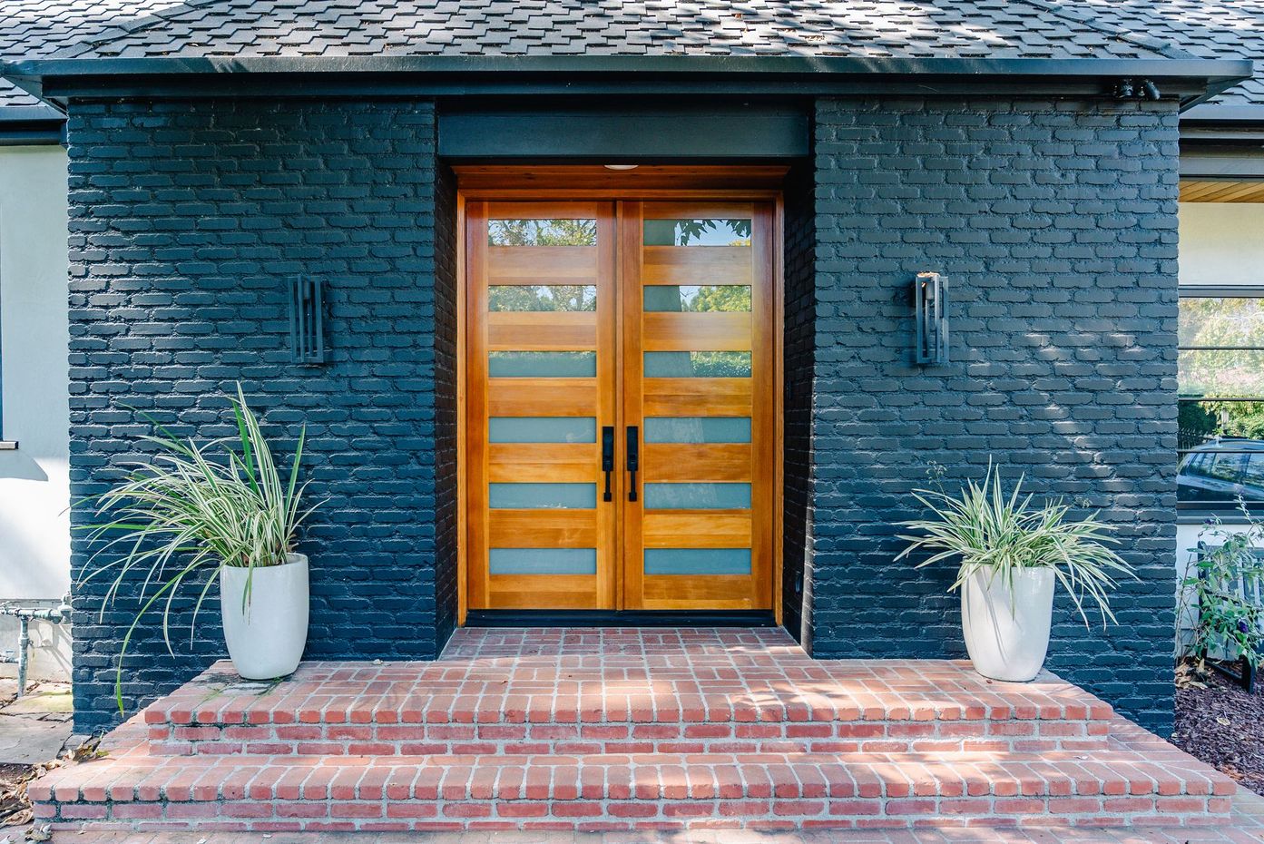 Front entrance with a wooden double door, dark brick facade, and two potted plants on a brick stoop.