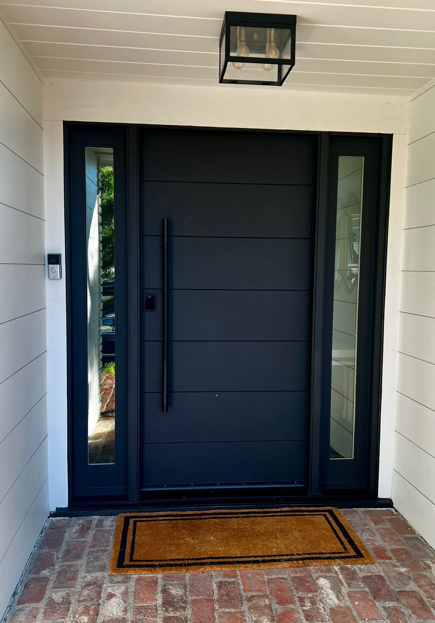 Modern black door with sidelights, black handle, welcome mat, brick steps.