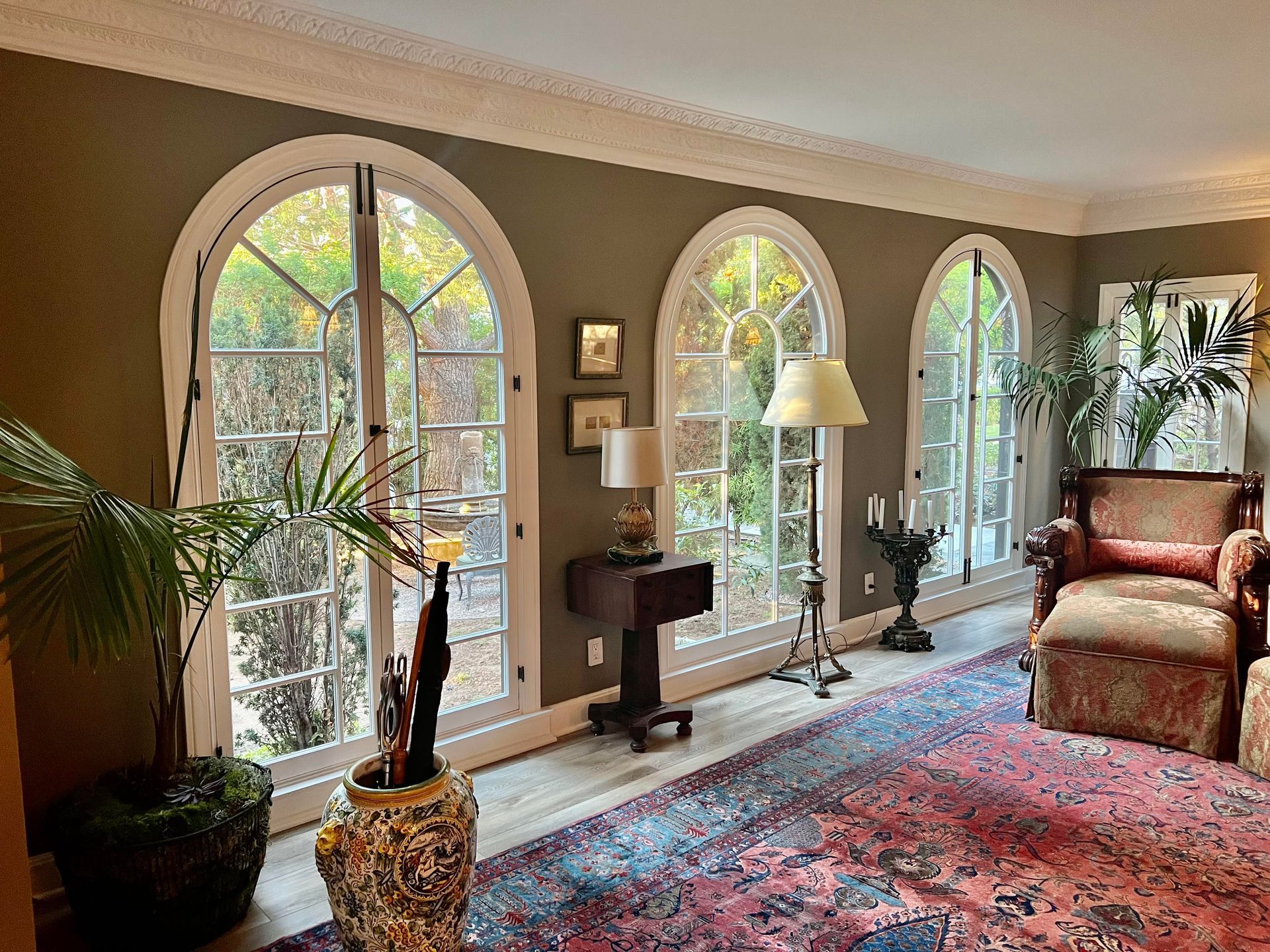 Living room with arched windows, olive-green walls, ornate rug, furniture, plants.