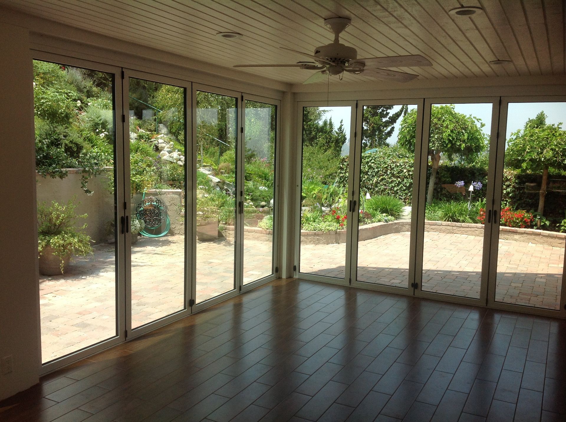 Bright room with large glass doors opening to a garden. Wooden floor, white ceiling with a fan.