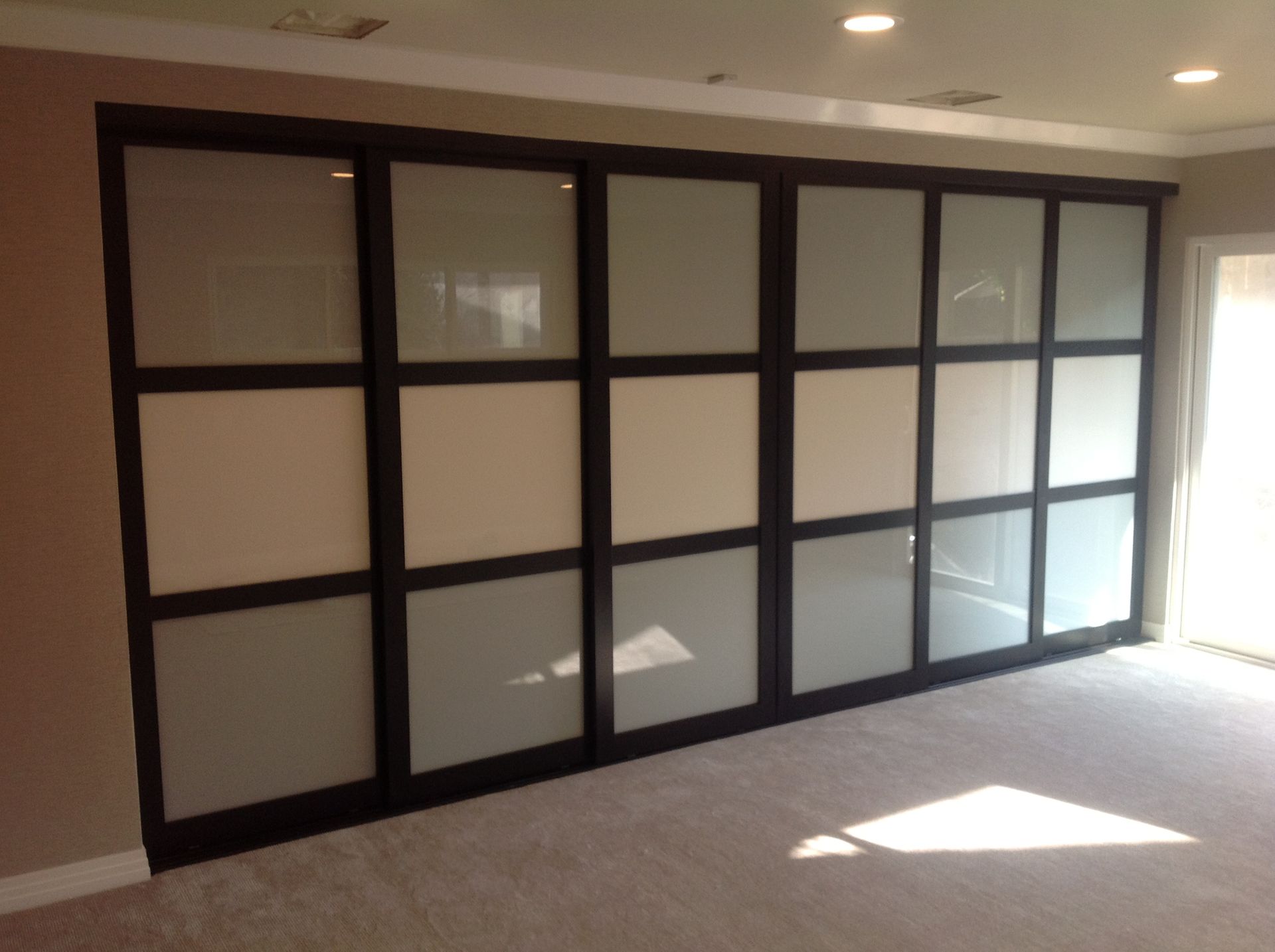 Sliding closet doors, dark frames, frosted glass panels, beige and gray, against a beige wall in a bright room.