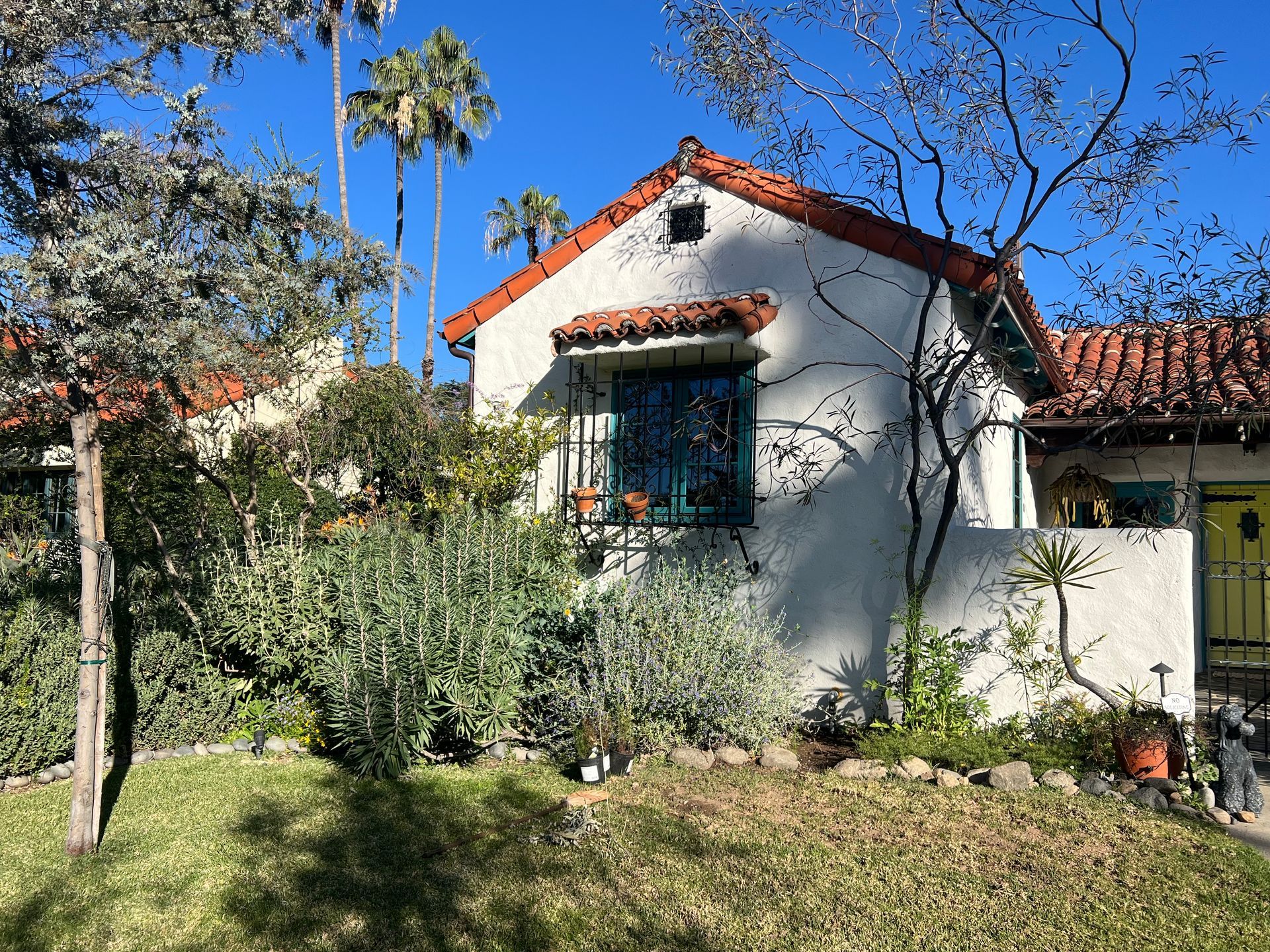 White stucco house with a red tile roof, blue window, and lush garden in bright sunlight