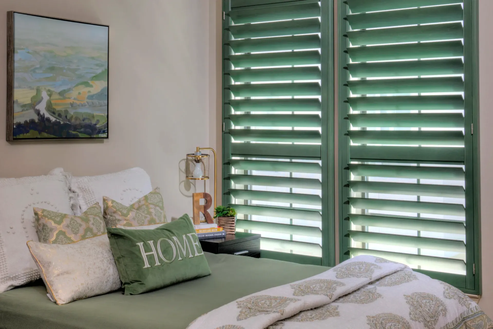 Green bedroom with a bed, white pillows, framed art, and teal shutters filtering light