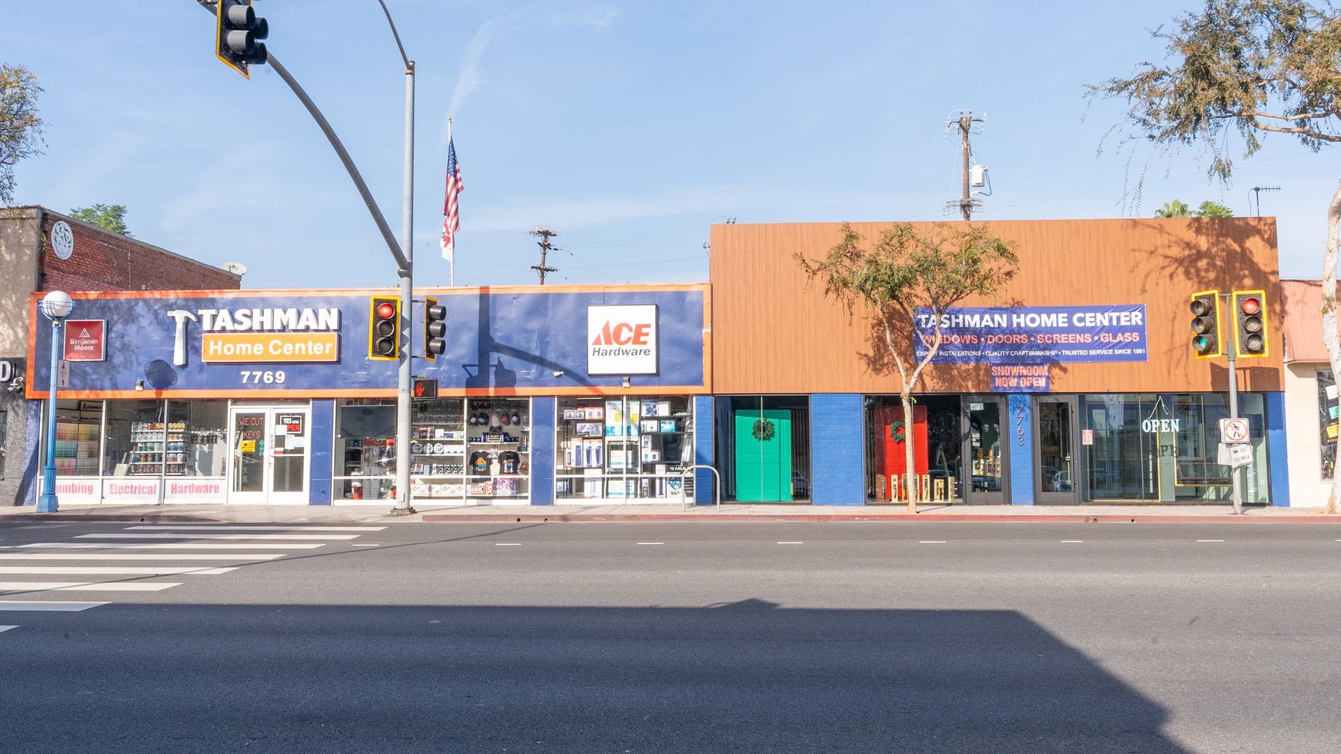 Buildings of T. Tashman, Ace Hardware, and an electrical repair shop on a sunny street, traffic lights overhead.