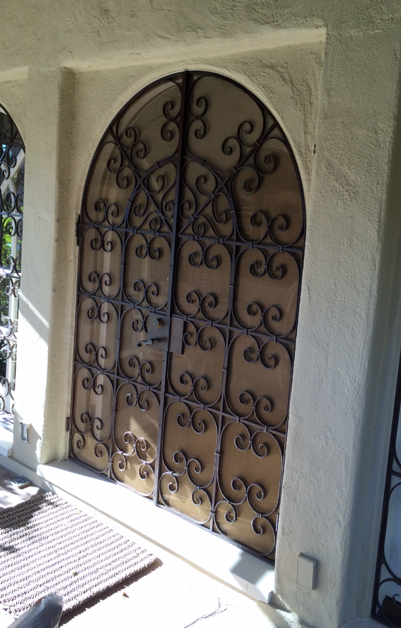 Decorative wrought-iron arched gate in a stucco wall beside a sunlit walkway